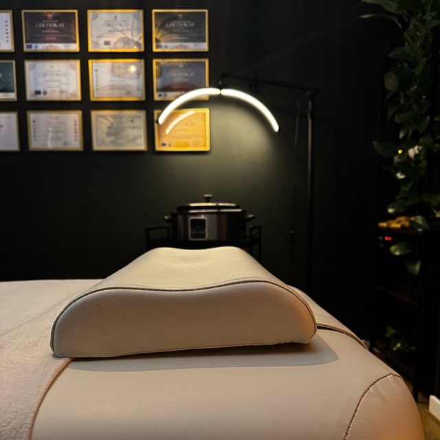 A massage treatment room with a neatly prepared massage table and a supportive face cradle in the foreground. A modern curved lamp provides focused, warm lighting above the table. In the background, framed certificates are displayed on the wall, highlighting professional qualifications, while a device for heating oils or stones and a plant add to the calm, professional atmosphere.
